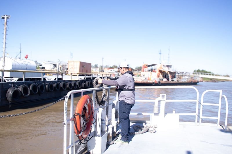 Port worker in a baseball/trucker hat and life vest handles heavy mooring rope on a vessel or dock on a sunny day.
