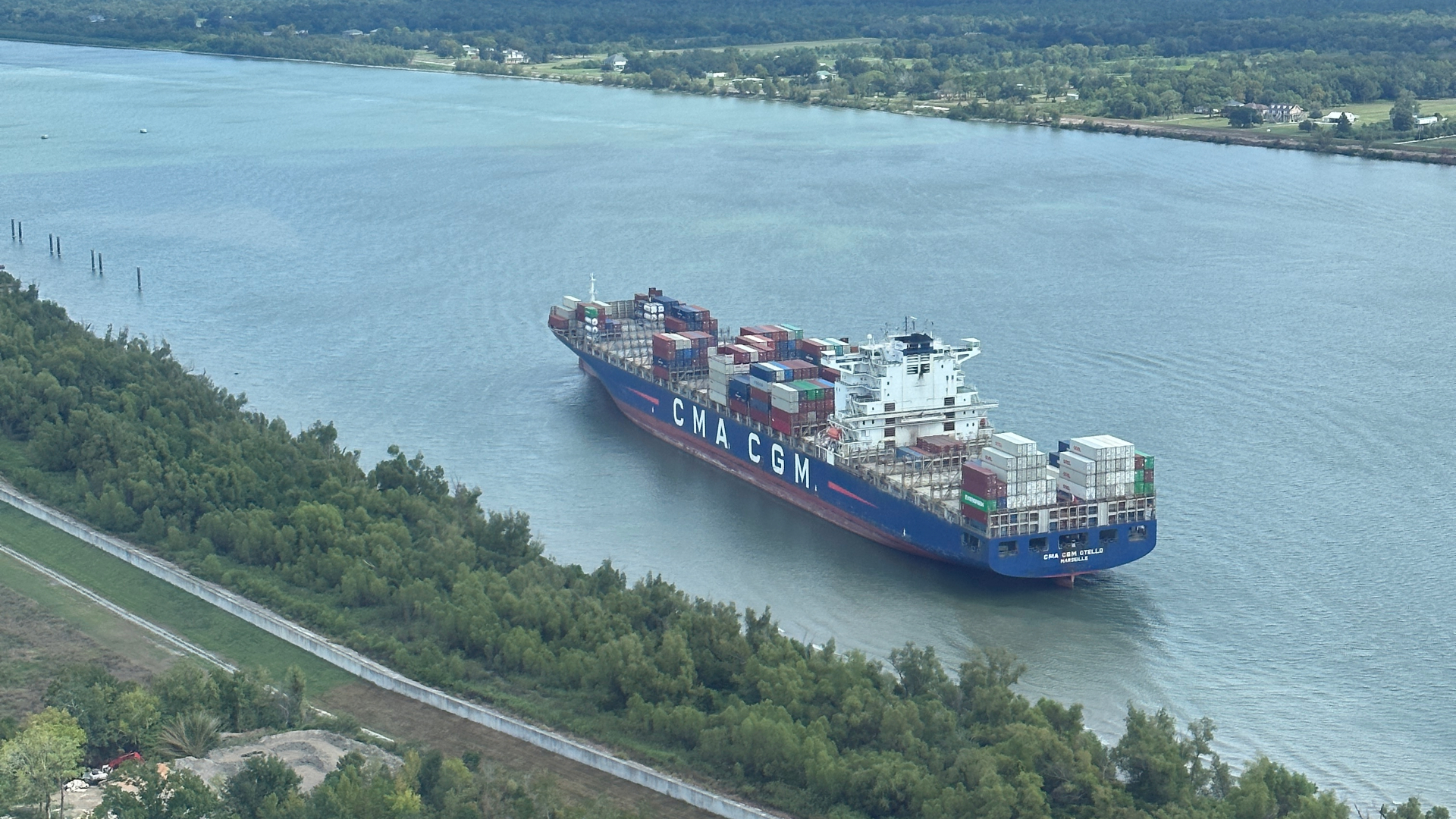 Aerial view of a large blue and red CMA CGM container ship traveling along a wide river. The ship is loaded with multi-colored cargo containers. The river is flanked by green, wooded banks and a paved road or levee on the left side.