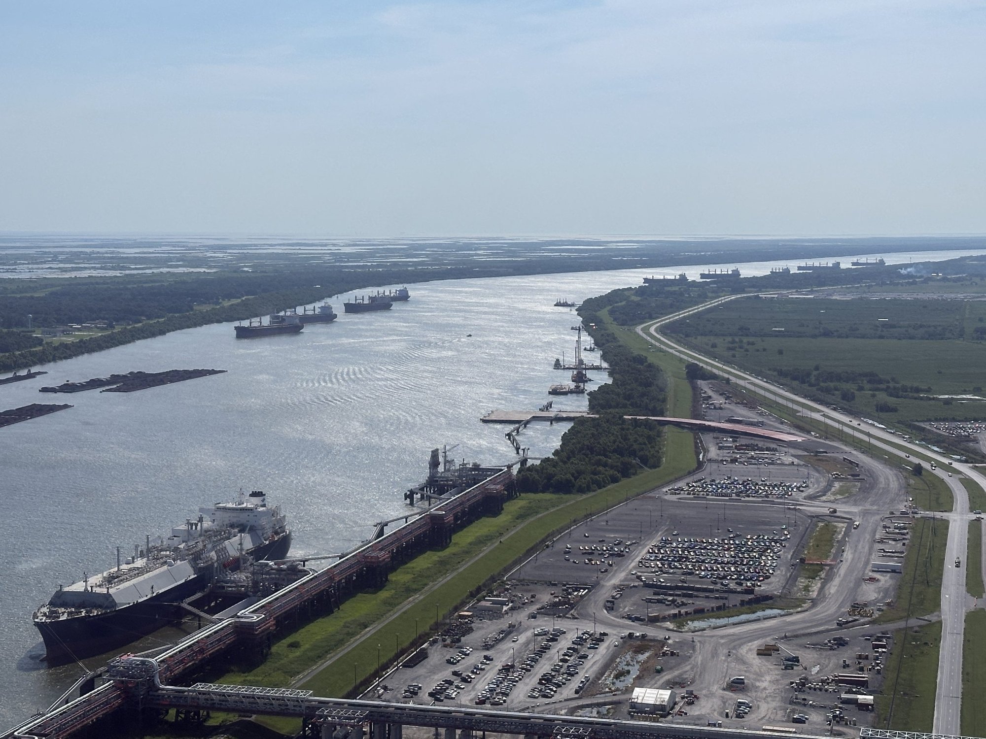 Wide aerial view of a large tanker docked at a terminal, with several other ships anchored further down a long, sunlit river