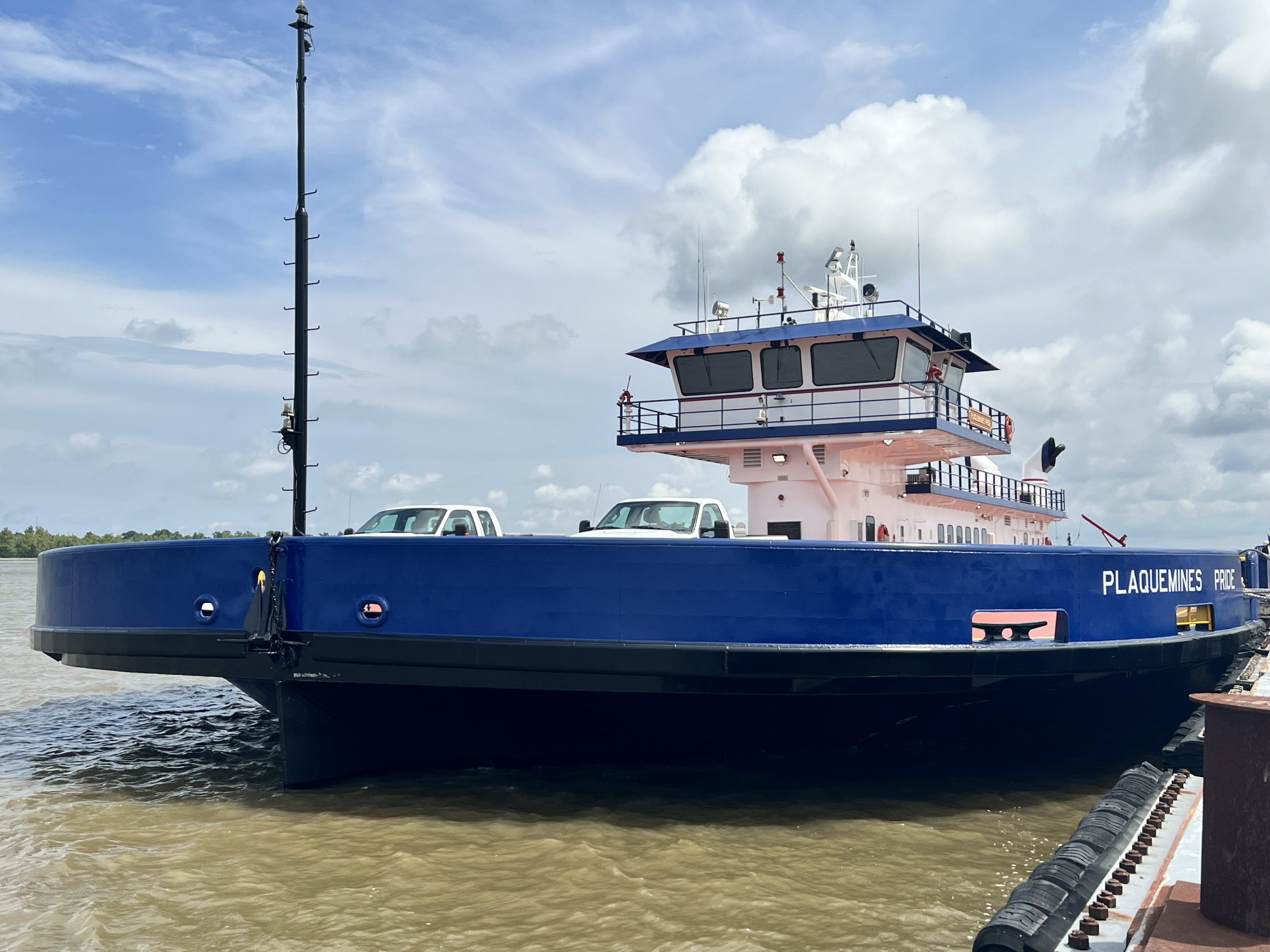 A blue and white ferry named "Plaquemines Pride" docked at a pier, carrying two white pickup trucks on its deck under a cloudy sky.