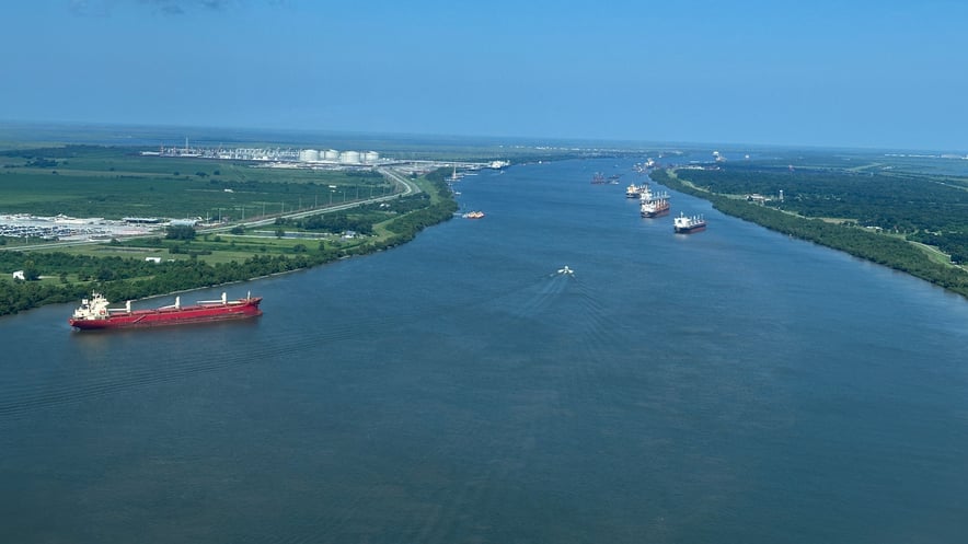 A wide, high-altitude aerial view of several large cargo vessels navigating a long stretch of the Mississippi River, with a vast white industrial facility visible on the left bank and dense green forests on the right.