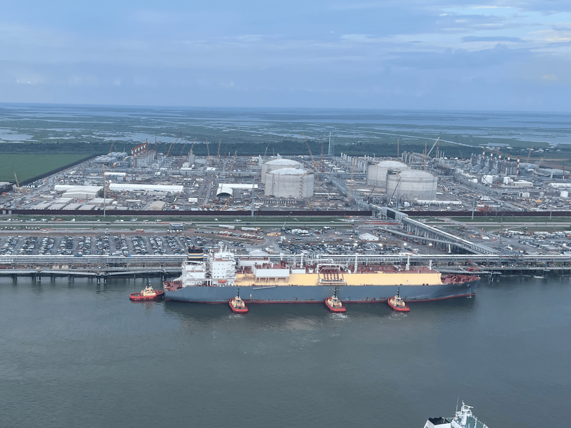 Aerial view of four red tugboats assisting a large gray and tan tanker as it maneuvers at a terminal adjacent to a massive industrial facility.