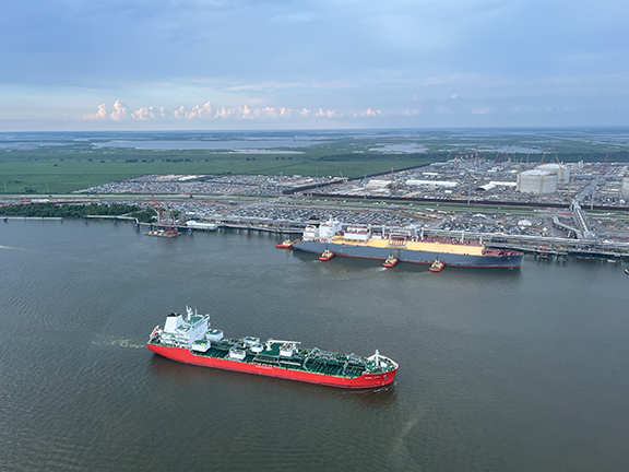 Aerial view of a wide river featuring two large vessels. In the foreground, a red and white tanker sails downstream. In the background, a massive grey and yellow LNG carrier is docked at an industrial port terminal, assisted by four red tugboats. The landscape is flat marshland under a hazy sky.